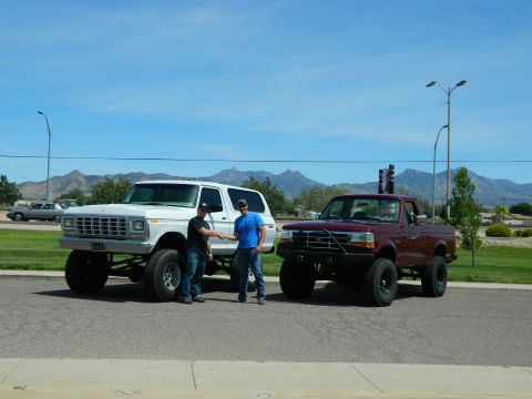 1979 Ford Bronco - Two men shake hands in front of a lifted white 1979 Ford Bronco and a lifted maroon Ford pickup truck parked on an asphalt road with mounta…