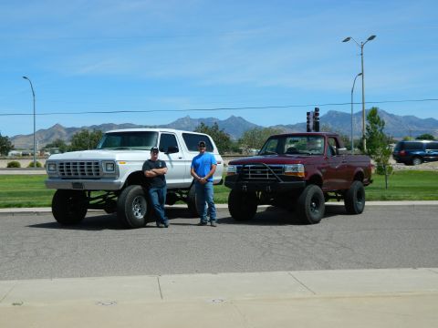1979 Ford Bronco - Two men stand next to a lifted white SUV and a lifted red pickup truck parked on an asphalt road with mountains in the background.