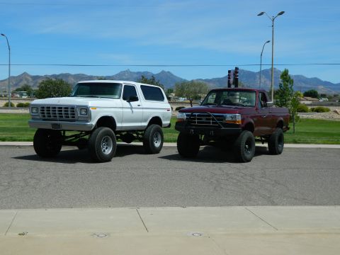 1979 Ford Bronco - A lifted white 1979 Ford Bronco is parked next to a lifted maroon Ford F-150 pickup truck on an asphalt road with mountains in the backgrou…