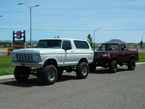 1979 Ford Bronco - A lifted white 1979 Ford Bronco is parked on asphalt next to a lifted maroon Ford pickup truck. A Route 66 sign is visible in the backgroun…
