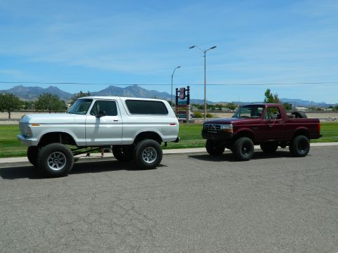 1979 Ford Bronco - A lifted white 1979 Ford Bronco is parked on asphalt next to a lifted maroon Ford Bronco pickup truck. Both vehicles are facing right under…