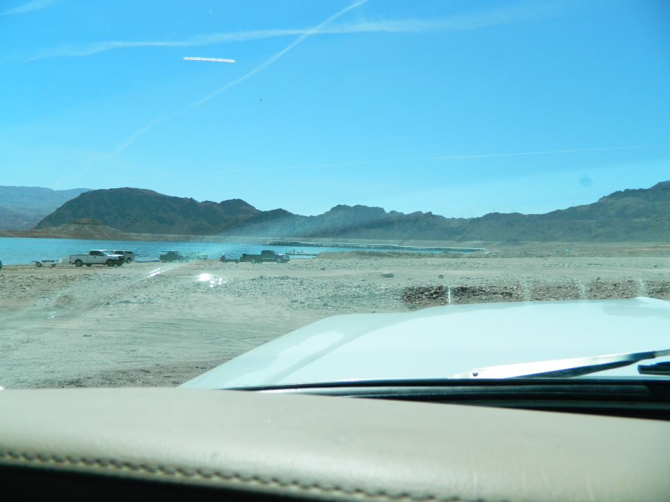 View from inside a vehicle looking out at a desert lake with mountains and parked trucks.