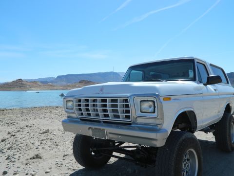1979 Ford Bronco - A lifted white 1979 Ford Bronco is parked on a rocky shore next to a bright blue lake. The front of the vehicle is angled towards the camer…
