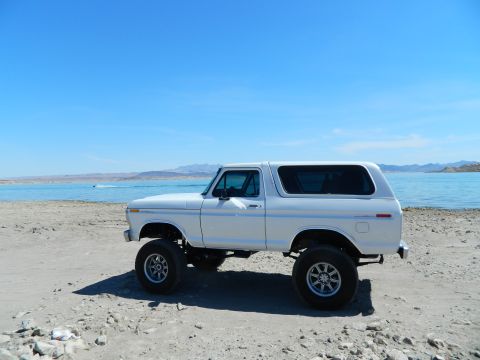 1979 Ford Bronco - A lifted white 1979 Ford Bronco is parked on a sandy beach next to a body of water under a clear blue sky. The vehicle is viewed from the s…