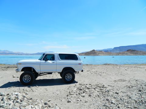 1979 Ford Bronco - A lifted white 1979 Ford Bronco is parked on a rocky shore next to a bright blue lake with mountains in the background.