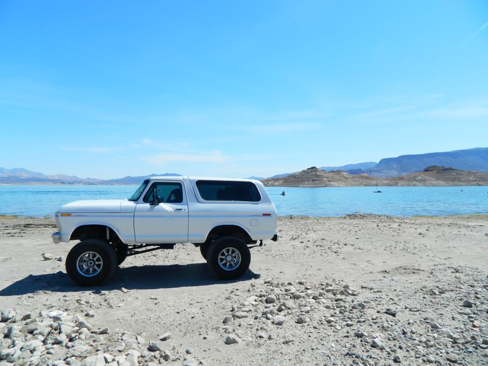 White 1979 Ford Bronco parked on a rocky shore by a blue lake.