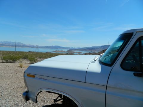 1979 Ford Bronco - The front right quarter of a white 1970s Ford pickup truck is shown parked on a gravelly surface with a large body of water and mountains i…