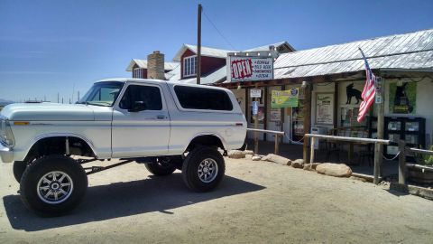 1979 Ford Bronco - A lifted, white 1979 Ford Bronco is parked in front of a rustic general store with an American flag flying. The vehicle has large off-road…