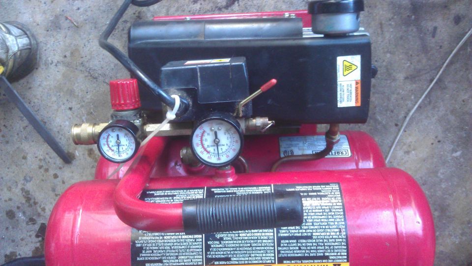 Red air compressor with gauges and black motor housing on a wet concrete surface.