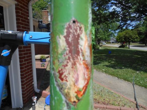 Damaged green pole with rust in foreground, blue bicycle repair stand and brick house in background.
