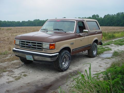 1987 Ford Bronco - A two-tone brown 1987 Ford Bronco is parked on a dirt road with fields in the background.