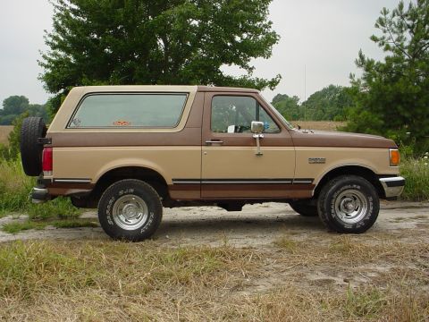 1987 Ford Bronco - This two-tone brown Ford Bronco is shown from the side, parked outdoors on a dirt road with grassy areas and trees.