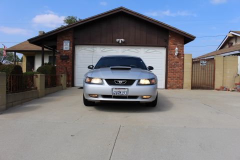 2002 Ford Mustang - The front view of a silver Ford Mustang parked in a driveway in front of a garage. The car has its headlights and fog lights on.