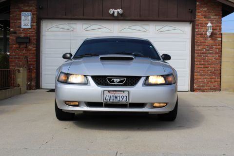 2002 Ford Mustang - Front view of a silver Ford Mustang parked in front of a garage. The car has its headlights and fog lights on, and a California license pla…