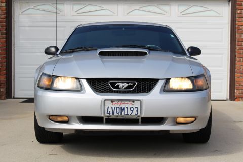 2002 Ford Mustang - Front view of a silver Ford Mustang parked in front of a white garage door. The car has its headlights on and displays a California license…