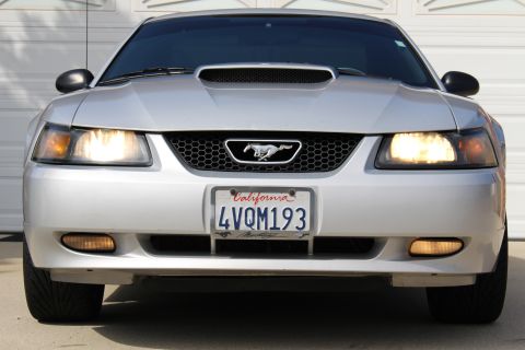 2002 Ford Mustang - Front view of a silver Ford Mustang with its headlights on, parked in front of a garage door. The California license plate reads "4VQM193".