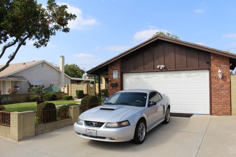 2002 Ford Mustang - A silver Ford Mustang coupe is parked in a driveway in front of a brick and wood-sided garage on a sunny day. The car has a license plate t…