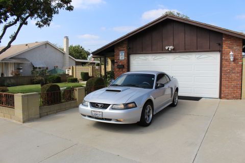 2002 Ford Mustang - A silver Ford Mustang coupe is parked in a driveway in front of a brick and wood-sided garage. The car is facing slightly towards the viewe…