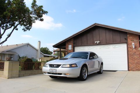 2002 Ford Mustang - A silver Ford Mustang coupe is parked in a driveway in front of a brick and wood-sided garage. The car is facing the camera at a three-quar…