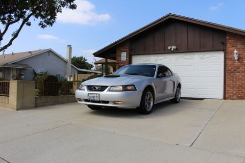 2002 Ford Mustang - A silver 2002 Ford Mustang coupe is parked in a driveway in front of a brick and wood-sided garage. The car is facing slightly left, with i…