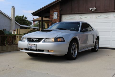2002 Ford Mustang - A silver 2002 Ford Mustang GT coupe is parked in a driveway in front of a garage. The car has a black grille with a Mustang emblem and a Ca…