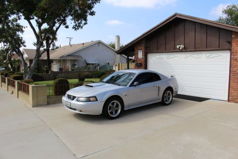 2002 Ford Mustang - A silver 2002 Ford Mustang GT coupe is parked in a driveway in front of a suburban garage. The car is viewed from a three-quarter front ang…
