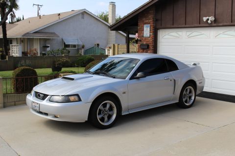 2002 Ford Mustang - A silver 2002 Ford Mustang GT coupe is parked in a driveway in front of a brick and wood garage. The car is viewed from a 3/4 front angle,…