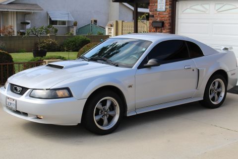 2002 Ford Mustang - A silver 2002 Ford Mustang GT coupe is parked in a driveway in front of a brick house with a white garage door. The car is viewed from a 3/…