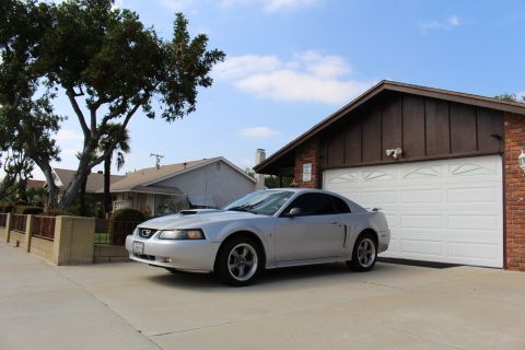 2002 Ford Mustang - A silver 2002 Ford Mustang coupe is parked in a driveway in front of a brick and wood-paneled garage. The car is viewed from a 3/4 front an…