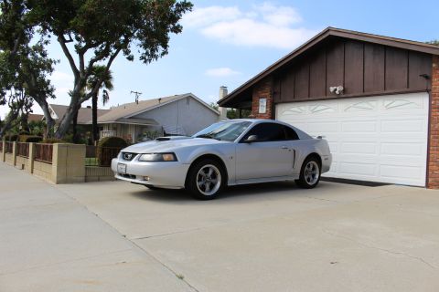 2002 Ford Mustang - A silver Ford Mustang coupe is parked on a concrete driveway in front of a two-car garage with a white door. The car is angled towards the…