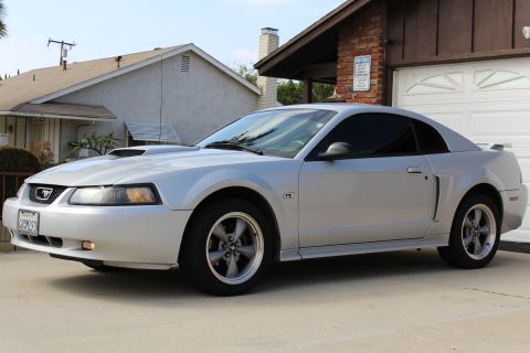 2002 Ford Mustang - A silver 2002 Ford Mustang GT coupe is parked in a driveway in front of a brick and wood garage. The car is viewed from the front three-qua…