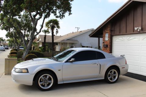 2002 Ford Mustang - A silver Ford Mustang GT coupe is parked in a driveway next to a brick and wood-sided garage. The car is shown from the side, facing right,…
