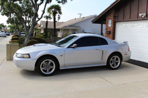 2002 Ford Mustang - A silver 2002 Ford Mustang GT coupe is parked in a driveway next to a garage. The car is viewed from the side, showing its tinted windows a…