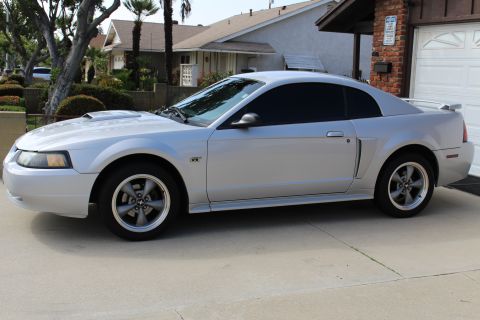 2002 Ford Mustang - The side profile of a silver Ford Mustang GT coupe is parked on a concrete driveway in front of a residential garage. The car features tint…