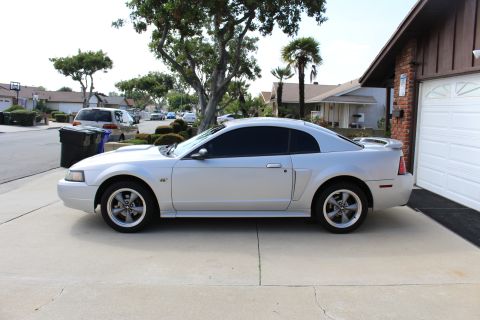 2002 Ford Mustang - The side profile of a silver Ford Mustang coupe is parked in a driveway next to a house with a white garage door. The car has tinted window…