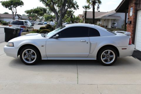 2002 Ford Mustang - The side profile of a silver 2002 Ford Mustang GT coupe is parked on a concrete driveway in front of a brick garage.