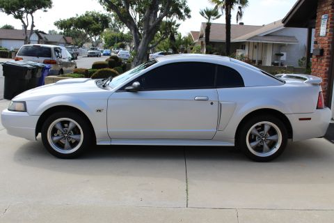 2002 Ford Mustang - The side view of a silver Ford Mustang GT coupe is parked on a concrete driveway in front of a residential house. The car has tinted window…