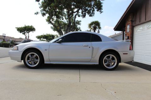 2002 Ford Mustang - Side profile of a silver 2002 Ford Mustang GT coupe parked on a concrete driveway in front of a garage. The car has tinted windows and afte…