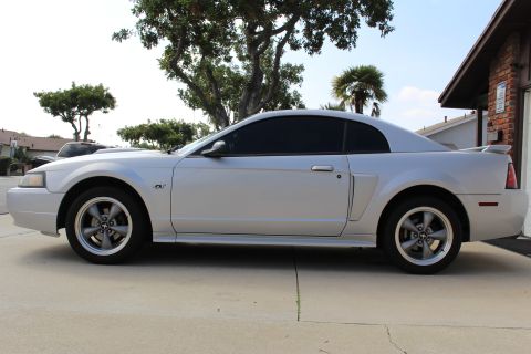 2002 Ford Mustang - Side profile of a silver 2002 Ford Mustang GT coupe parked on a concrete driveway. The car features tinted windows, a rear spoiler, and fiv…