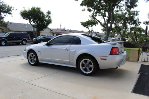 2002 Ford Mustang - The side profile of a silver Ford Mustang GT coupe is parked on a concrete driveway. A black SUV and a dark sedan are parked on the street…