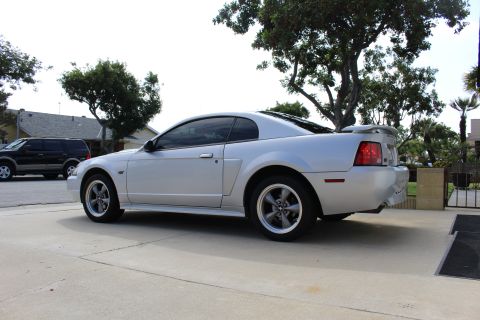 2002 Ford Mustang - The side profile of a silver Ford Mustang coupe is shown parked on a concrete driveway. A black SUV is parked behind it in the background.
