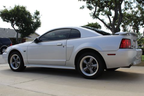 2002 Ford Mustang - The side profile of a silver Ford Mustang GT coupe is shown parked on a concrete driveway. The car features a rear spoiler and aftermarket…