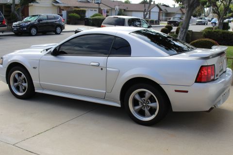 2002 Ford Mustang - The rear quarter view of a silver Ford Mustang coupe is shown parked on a concrete driveway. The car features tinted windows, a rear spoile…