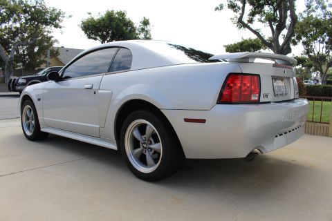 2002 Ford Mustang - The rear passenger side of a silver 2002 Ford Mustang GT coupe is shown parked on a concrete driveway. The car features a rear spoiler, aft…