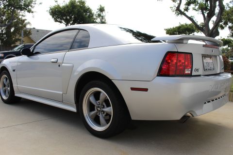 2002 Ford Mustang - The rear passenger side quarter of a silver Ford Mustang GT coupe is shown parked on a concrete driveway. The car features a rear spoiler,…