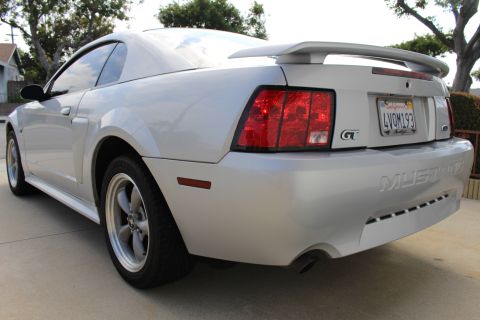 2002 Ford Mustang - The rear passenger side quarter of a silver Ford Mustang GT is shown parked on a concrete driveway. The car features a rear spoiler, tinted…