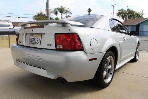2002 Ford Mustang - Rear 3/4 view of a silver Ford Mustang GT coupe parked on a concrete driveway. The car features a rear spoiler, custom license plate, and a…