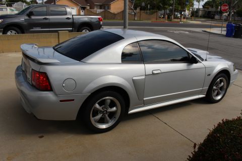 2002 Ford Mustang - The rear passenger side view of a silver 2002 Ford Mustang GT coupe is shown parked on a concrete driveway. A dark gray Toyota Tundra picku…