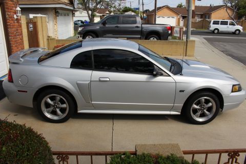 2002 Ford Mustang - The side profile of a silver Ford Mustang coupe is parked on a concrete driveway in front of a house. A dark gray pickup truck is parked be…
