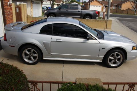 2002 Ford Mustang - The side view of a silver 2002 Ford Mustang GT coupe is parked in a driveway. A dark grey pickup truck is parked behind it in the same driv…